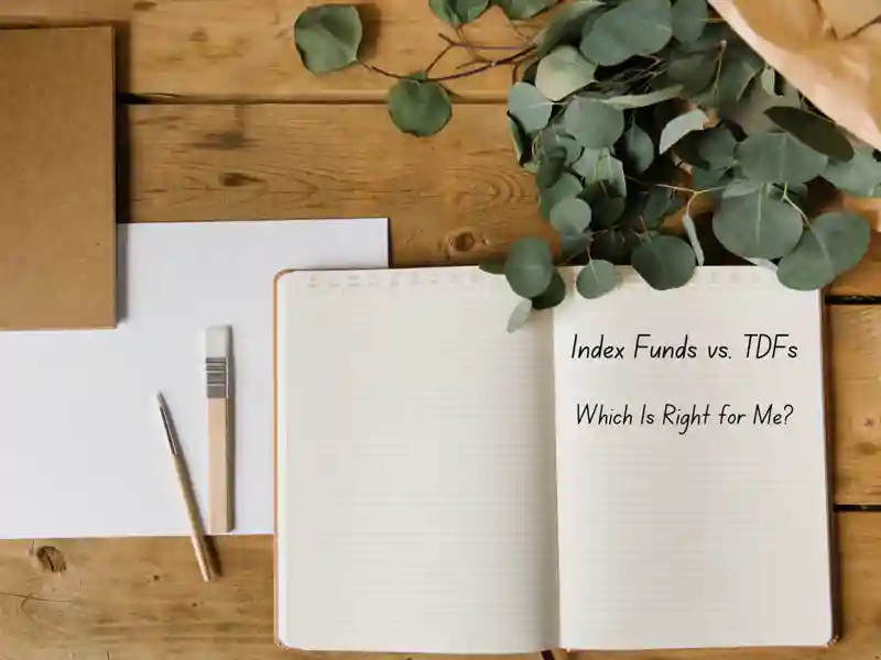 A notebook with "Index Funds vs. TDFs" written on it, surrounded by paint brushes, paper, and plants on a wooden desk.