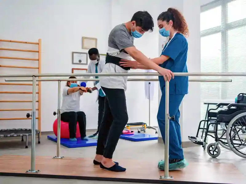 Young man using parallel bars to walk and physiotherapist helping him while another patient and professional are walking at the background.