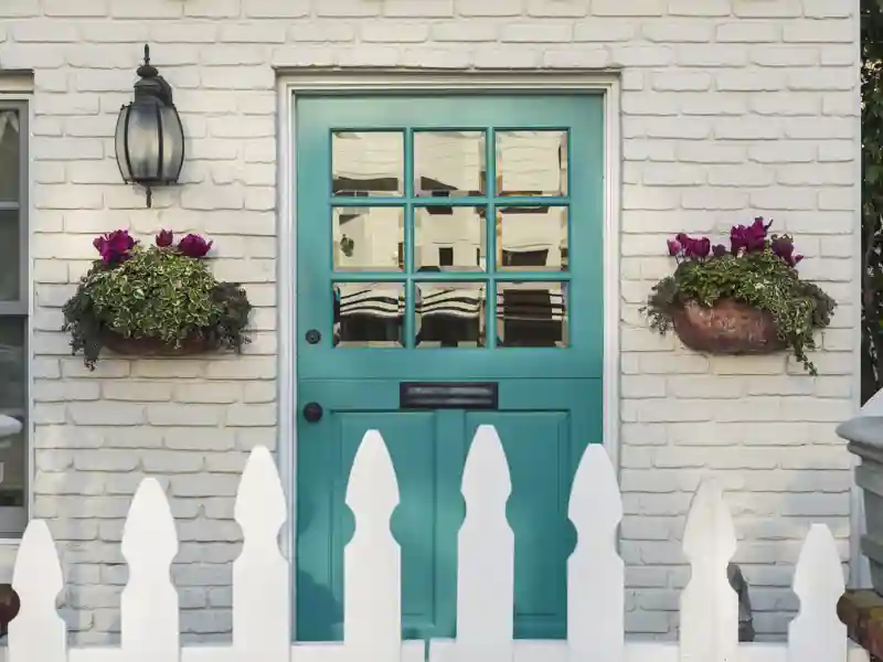 A teal wooden front door to a home, with white picket fence gate in foreground.