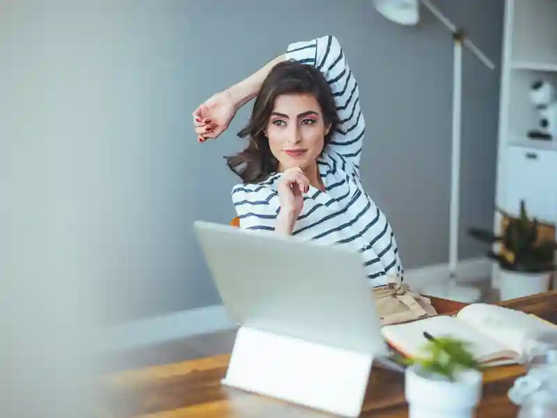A woman stretches at her desk while looking at a laptop, with books and plants nearby.