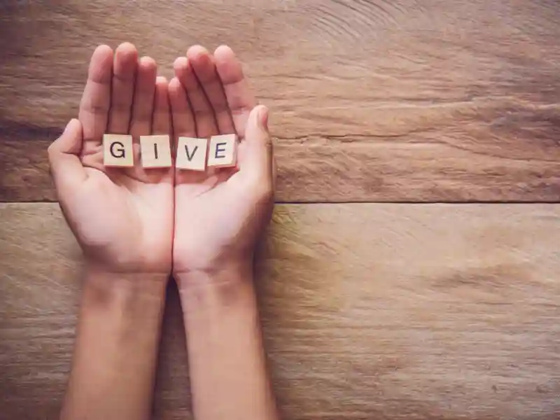 Hands holding wooden blocks that spell the word give, positioned on a wooden table.