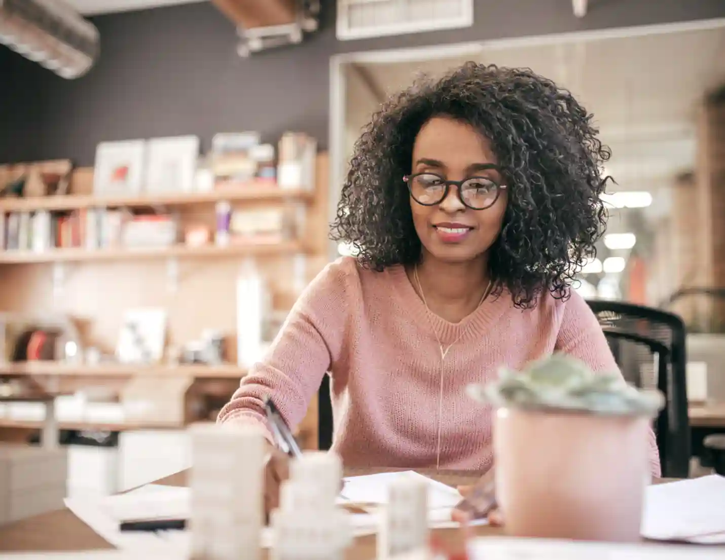 A woman wearing glasses is seated at a desk, surrounded by papers, focused on her work.